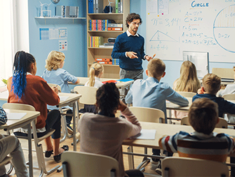 Elementary Classroom of Diverse Bright Children Listening Attentively to their male teacher standing at the top of the classroom.